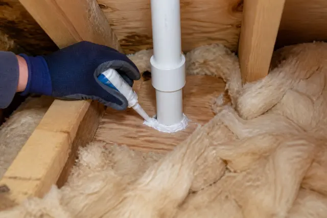 Gloved homeowner air-sealing a plumbing penetration in an attic floor.
